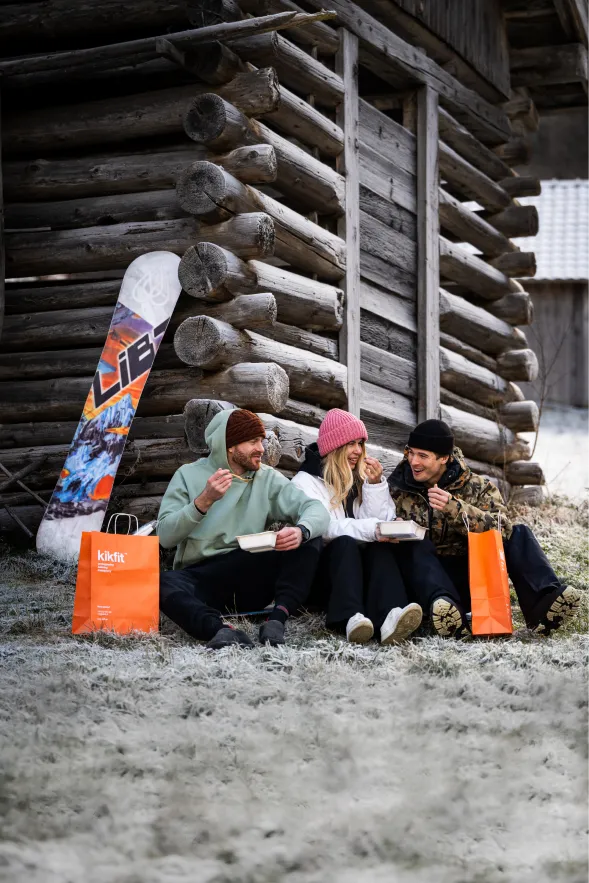 Trzy osoby jedzą na zewnątrz zimą przy drewnianej chacie, obok torby kikfit i snowboardu