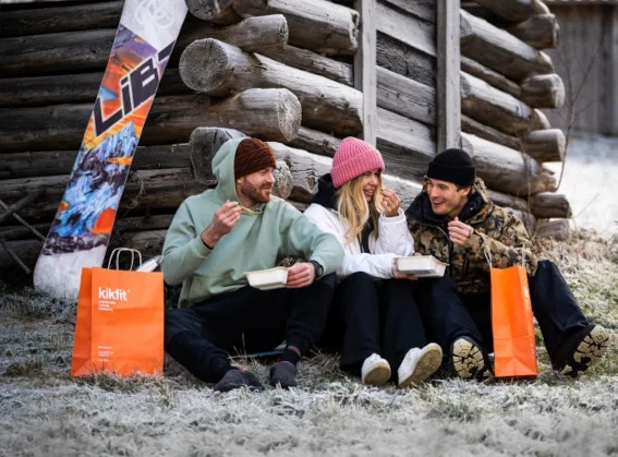 Trzy osoby jedzą na zewnątrz zimą przy drewnianej chacie, obok torby kikfit i snowboardu
